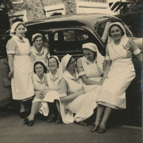 Zwart wit foto met groep van 7 verpleegsters in werk unifrom voor een kleine auto voor de kliniek van de kraamafdeling in Den Haag.