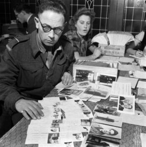 Zwart wit foto van op de voorgrond een man achter een tafel en achter hem aan dezelfde tafel een vrouw. De tafel ligt vol met documenten waaronder ook veel portretfoto's.